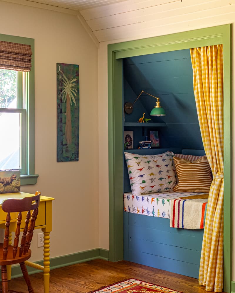 Book nook in a kid's room with a yellow gingham curtain, green reading lamp and colorful dinosaur sheets.