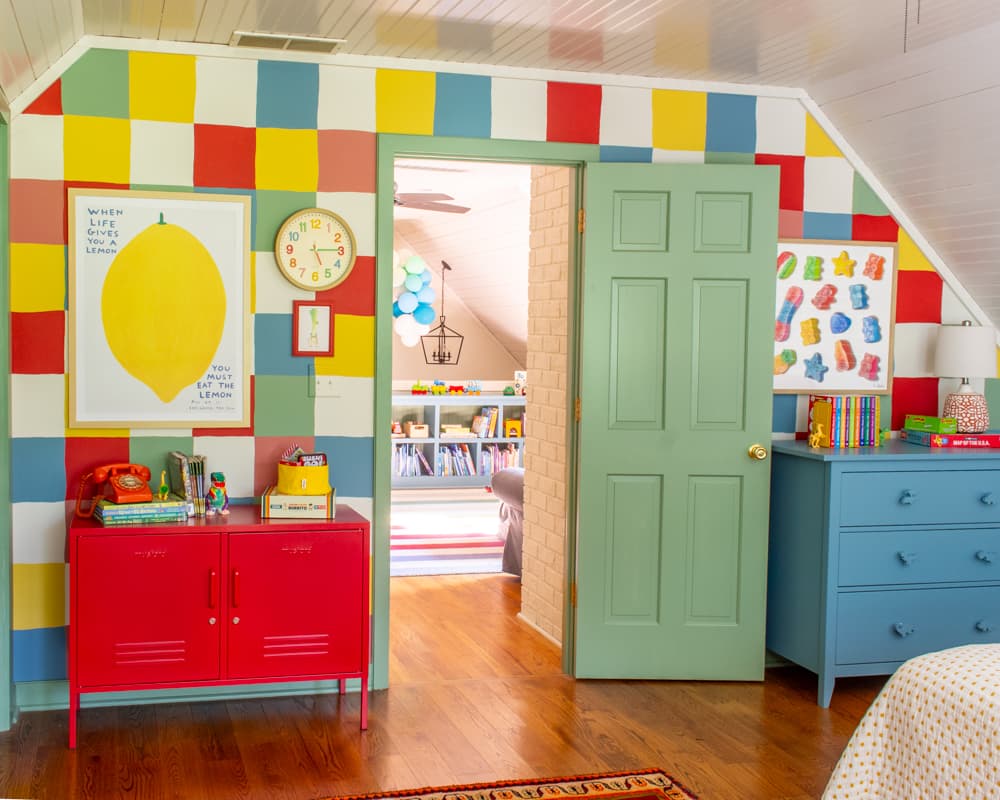 Colorful kids room with checkered wall, colorful art, blue dresser, green door and red cabinet.