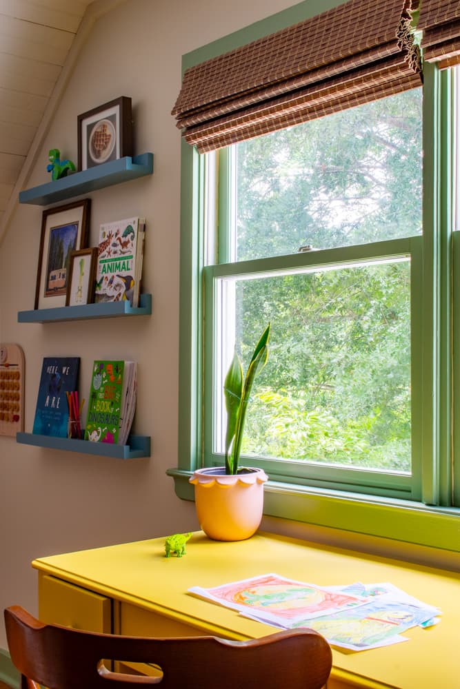 Yellow desk in a kids room under a window with green trim and next to blue ledge shelves.