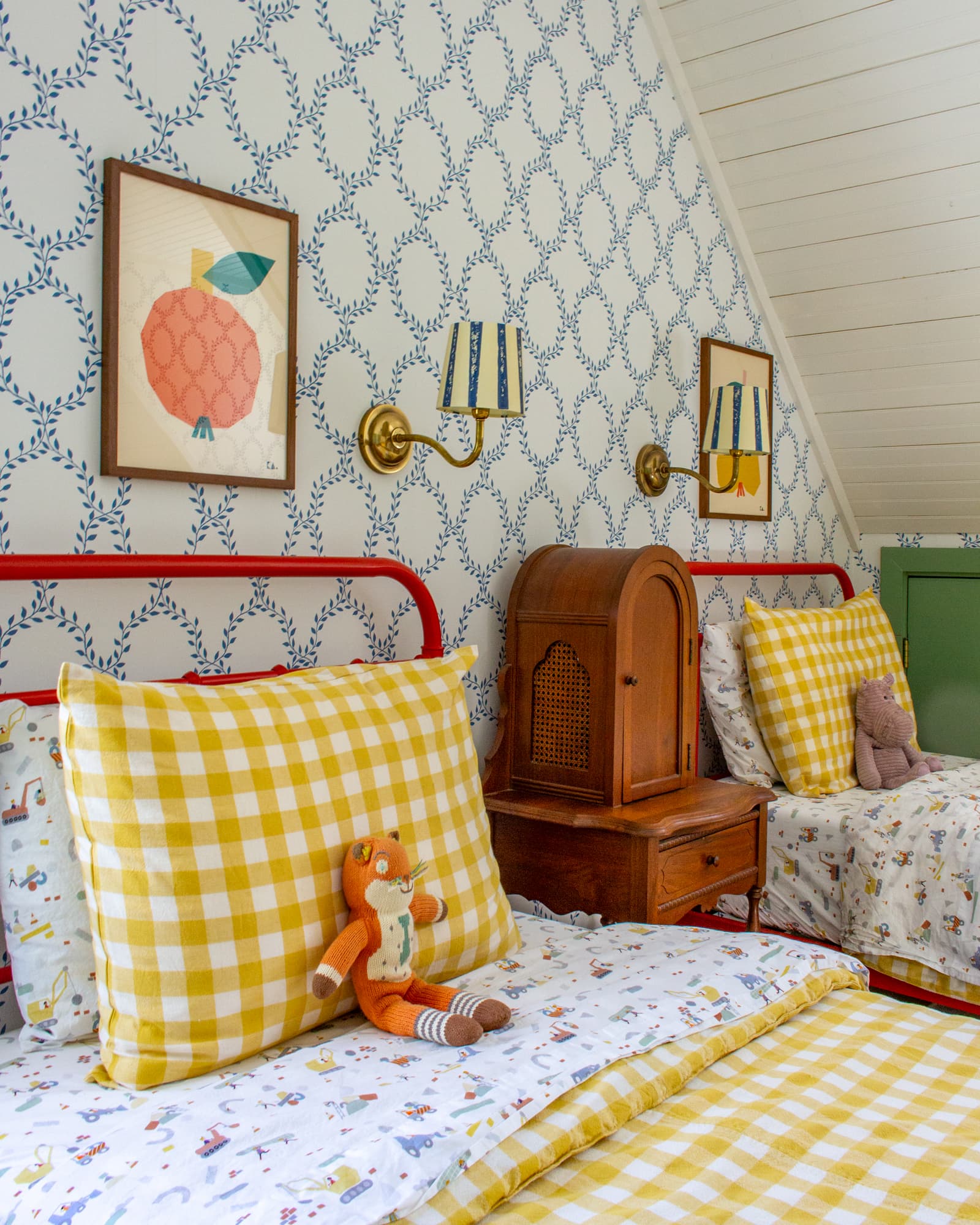 Red twin beds in a child's bedroom with colorful art above each. An antique phone cabinet sits between the two beds