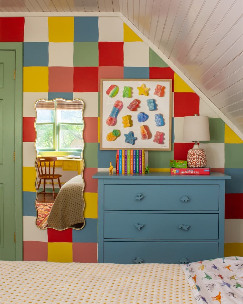 Kids room with a colorful checkered wall and a blue dresser, art print of gummy candy and a wavy framed mirror.
